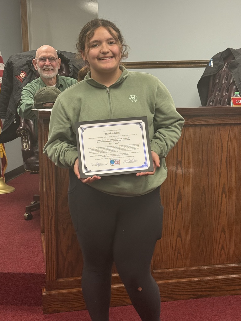 A student stands in a meeting room on red carpet, holding a framed certificate. The certificate recognizes Elizabeth Collins, Division II, Anthropology, Group, “Man O’ War.” The student is positioned in front of a wooden boardroom desk, with another individual seated behind the desk. An American flag is visible in the background.