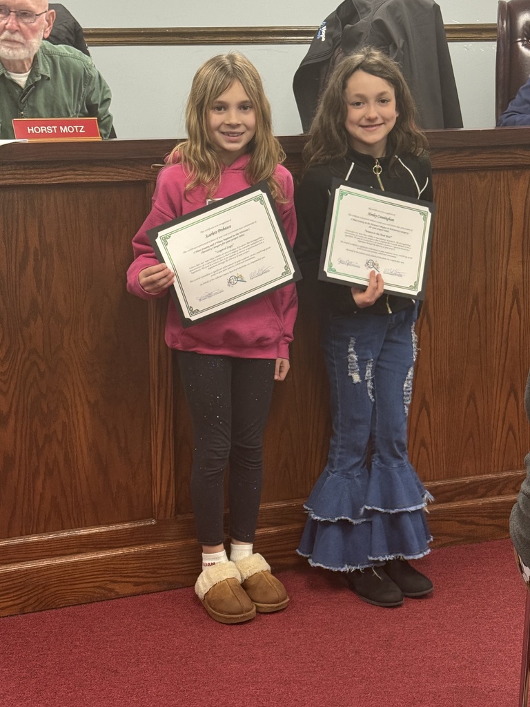 Two students stand side by side in front of a wooden boardroom desk, each holding a certificate. The student on the left is holding an award for Henley Cunningham – Division I, Physics and Astronomy, “Bounce to the Heat Beat.” The student on the right is holding an award for Scarlett Probasco – Division I, Chemistry I, “Layered Logic,” Regional 3rd Place. Both are standing on red carpet with the certificates displayed prominently.