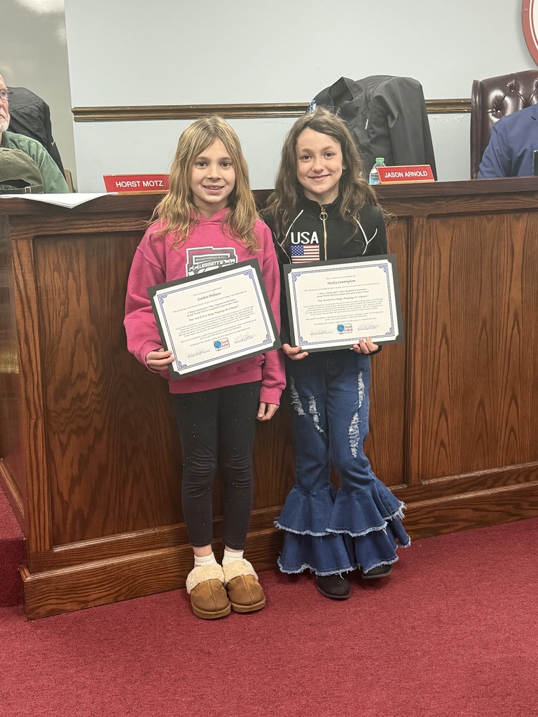 Two students stand side by side in front of a wooden boardroom desk, each holding a certificate. The student on the left holds an award for Henley Cunningham – Division I, World History, Group, “One World Five Rings: Mapping the Olympic.” The student on the right holds a matching certificate for Scarlett Probasco for the same project. Both students stand on red carpet, and nameplates and chairs from the meeting room are visible behind them.