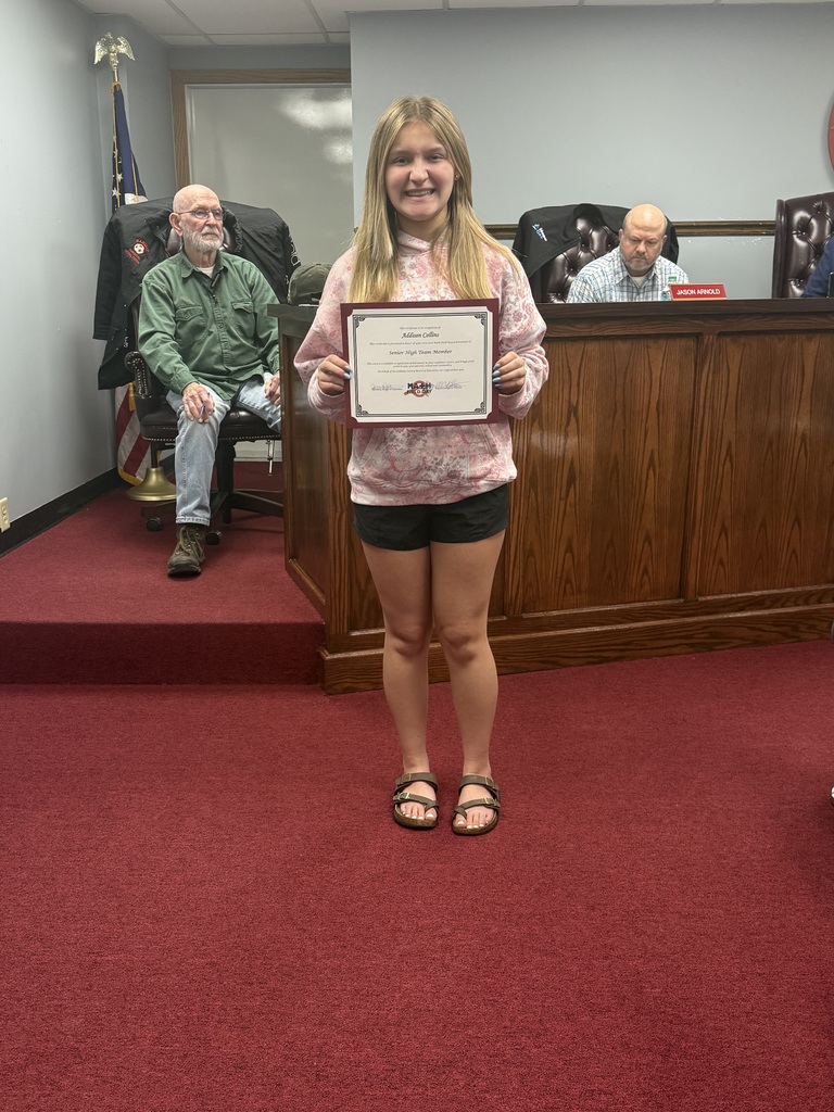 A student stands on red carpet in a meeting room holding a framed certificate recognizing Addison Collins for placing as a Senior High Team Member in Math Field Day. Behind the student is a wooden boardroom desk with seated individuals and an American flag visible near the corner of the room.