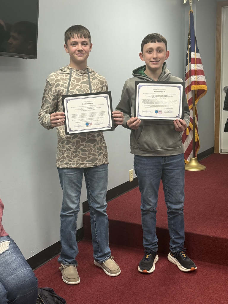 Two students stand side by side in a meeting room on red carpet, each holding a framed certificate. The student on the left holds an award recognizing participation in Division II, Economics, Group, “The Business Behind the Bar.” The student on the right holds a matching certificate for the same project. An American flag is visible in the background near a doorway.