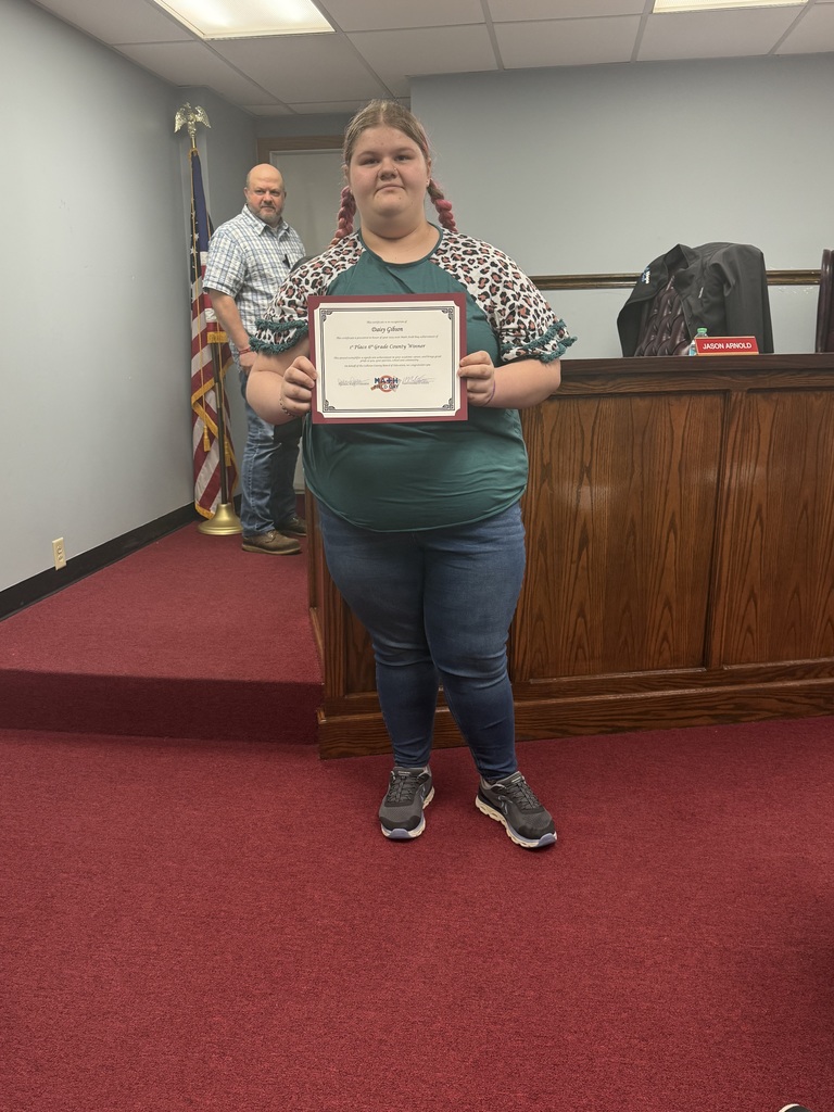 A student stands on red carpet in a meeting room holding a framed certificate. The certificate recognizes Daisy Gibson for earning 1st place in 6th Grade Math Field Day. A wooden boardroom desk and an American flag are visible in the background, along with another person standing near the wall.