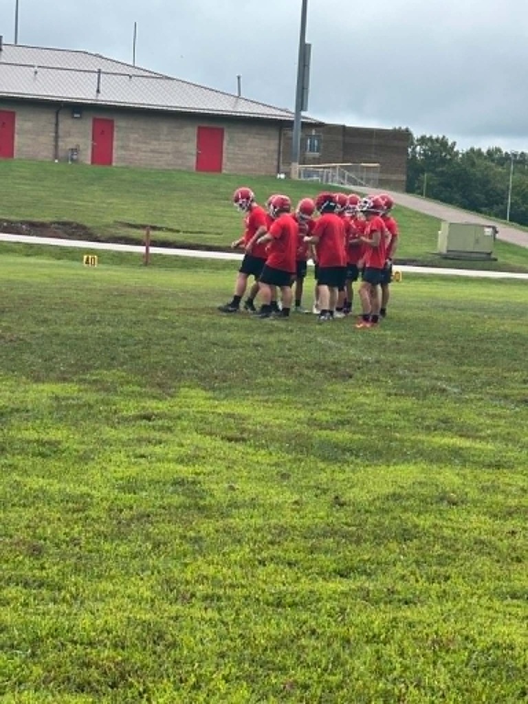 A group of young football players wearing red helmets and red shirts stand together in a huddle on a grassy football field. A light‑colored building with red doors sits in the background along with trees and a small hill.