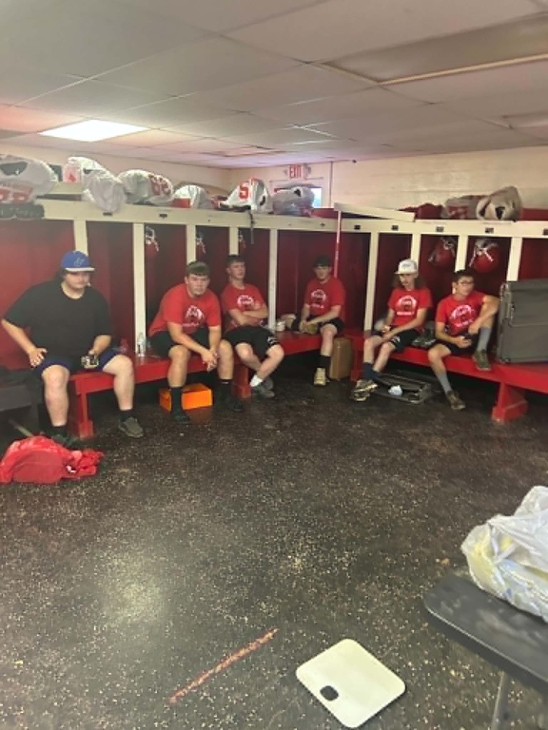 Six individuals wearing red shirts sit on a long red bench inside a football locker room. Open cubby‑style lockers behind them contain helmets and bags labeled with jersey numbers. Equipment bags and a small table are scattered around the room.