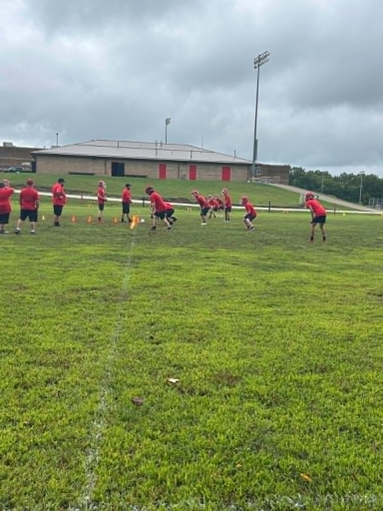 Football players in red shirts and black shorts practice on a grassy field with orange cones arranged in lines. A large brick building with red doors and tall light poles stands behind them under a cloudy sky.