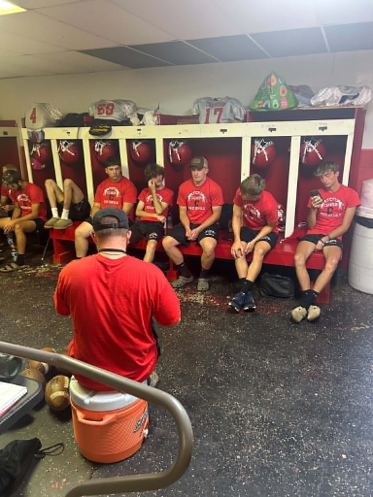 Several football players in red shirts and black shorts sit on a red bench in front of open lockers holding helmets and jerseys. A person sits on an orange water cooler facing the group, appearing to lead a discussion. The locker room has a speckled floor and some missing ceiling tiles.
