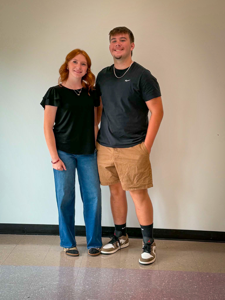 Two students stand side by side indoors against a plain light-colored wall for a posed school portrait. The student on the left is wearing a black short-sleeved shirt with ruffled sleeves, blue jeans, and dark shoes. The student on the right is wearing a dark athletic T-shirt, tan shorts, black socks, athletic shoes, and a necklace. Both are facing the camera and standing on a tiled floor with a black baseboard visible along the wall.