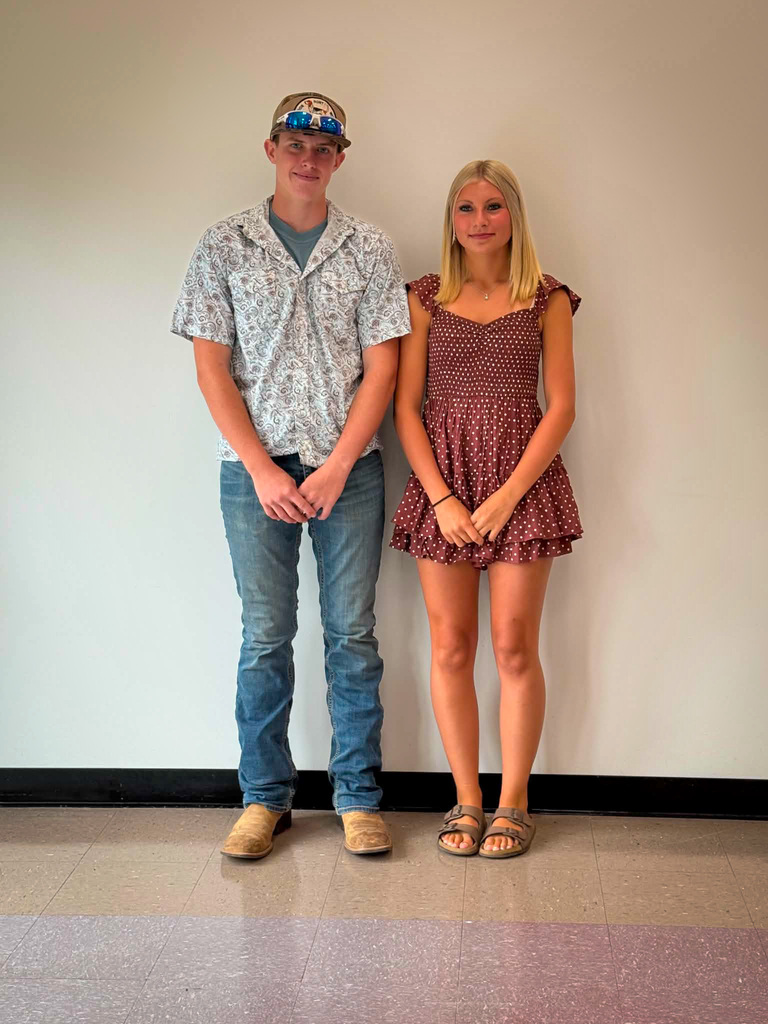 Two students stand side by side indoors against a plain light-colored wall for a posed school portrait. The student on the left is wearing a patterned short-sleeved button-up shirt over a blue T-shirt, blue jeans, tan boots, and a baseball cap with reflective sunglasses resting on the brim. The student on the right is wearing a maroon dress with a small white polka-dot pattern, ruffled sleeves, and sandals. Both are facing the camera with their hands clasped in front of them. The photo is taken on a tiled floor with a black baseboard visible along the wall.