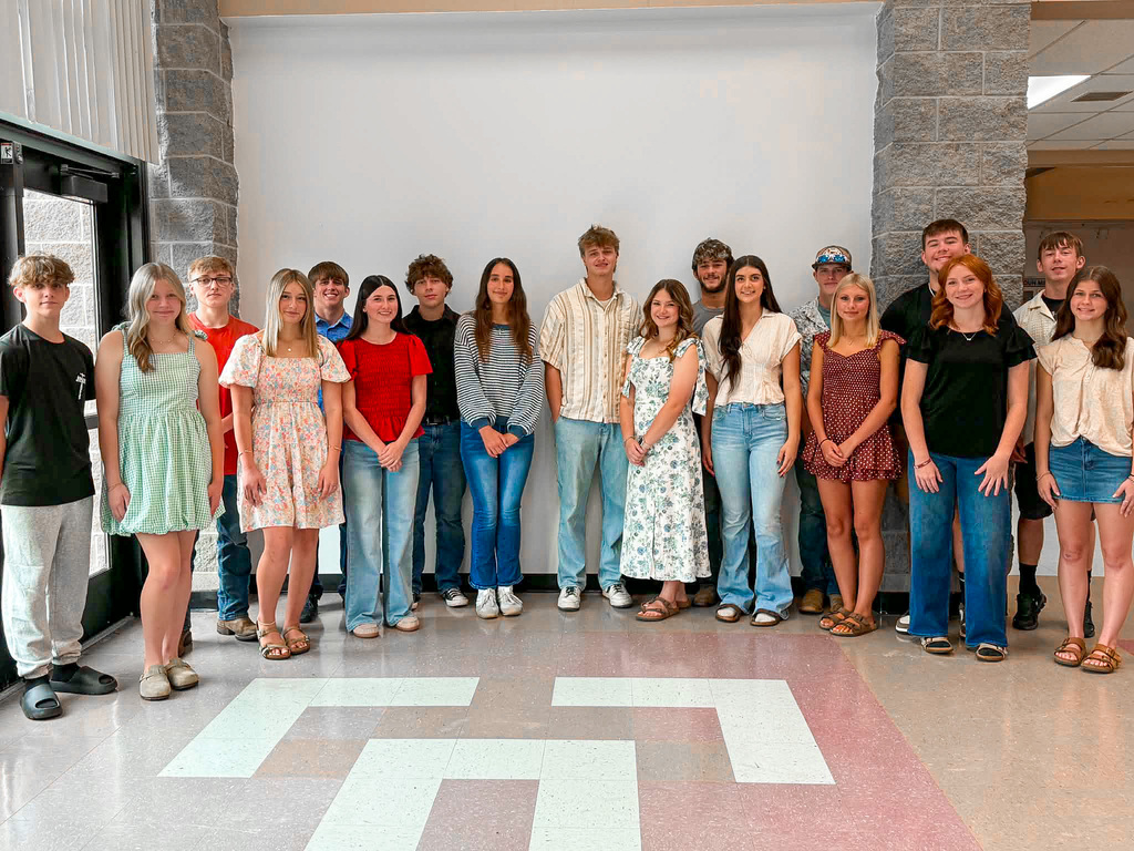 Group portrait of students standing indoors in a school hallway in front of a large white backdrop framed by gray brick walls. The group is arranged in a single row and includes students dressed in a variety of semi-formal and casual outfits, including dresses, blouses, jeans, button-up shirts, polo shirts, T-shirts, and sneakers. Several students wear light-colored floral or patterned clothing, while others wear solid colors such as red, black, blue, and white. The floor is made of multicolored tile, and a glass entrance door is visible on the left side of the image. The photo appears to be a formal group portrait of a school court or student representative group.
