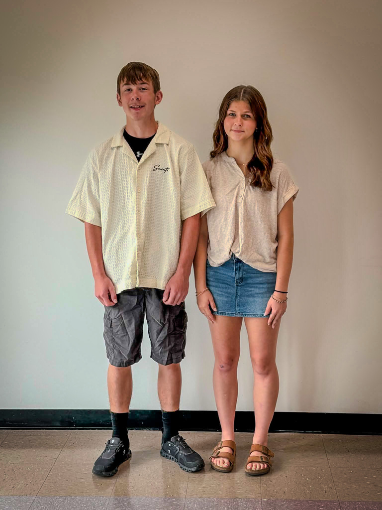 Two students stand side by side indoors against a plain light-colored wall. The student on the left is wearing a light short-sleeved button-up shirt over a dark shirt, dark cargo shorts, black socks, and dark athletic shoes. The student on the right is wearing a light short-sleeved blouse, a blue denim skirt, and brown sandals. Both are facing the camera with their arms at their sides. The floor is tiled, and the image appears to be a posed school portrait.