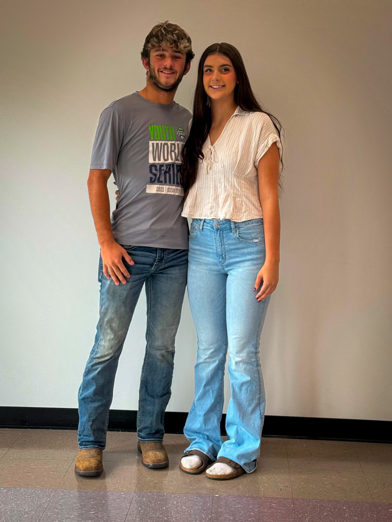 Two students stand side by side indoors against a plain light-colored wall for a posed school portrait. The student on the left is wearing a gray graphic T-shirt with blue and green lettering, blue jeans, and tan work boots. The student on the right is wearing a white short-sleeved blouse with subtle vertical stripes, light blue jeans, and light-colored sandals. Both are facing the camera and standing close together on a tiled floor, with a black baseboard visible along the wall behind them.