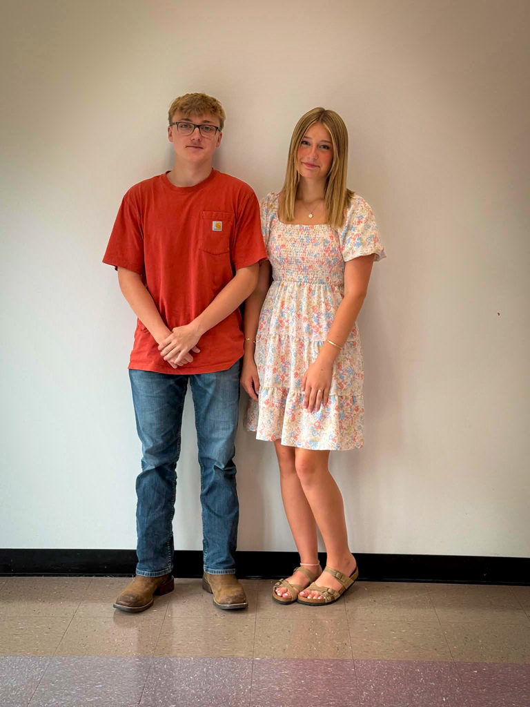Two students stand side by side indoors against a plain light-colored wall for a posed school portrait. The student on the left is wearing a red short-sleeved T-shirt, blue jeans, brown boots, and glasses, with hands clasped in front. The student on the right is wearing a light-colored floral dress with short sleeves and tan sandals. Both are facing the camera and standing on a tiled floor with a black baseboard visible along the wall.