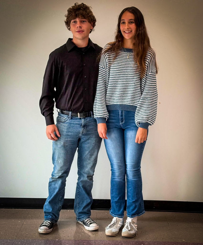 Two students stand side by side indoors against a plain light-colored wall for a posed school portrait. One student is wearing a dark long-sleeved button-up shirt, blue jeans, a black belt, and black-and-white sneakers. The other student is wearing a gray-and-white striped knit sweater with blue trim, blue jeans, a necklace, and white sneakers. Both are facing the camera and standing on a tiled floor, with a black baseboard visible along the wall behind them.