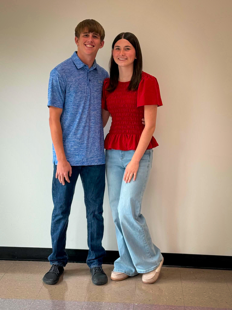 Two students stand side by side indoors against a plain light-colored wall for a posed school portrait. The student on the left is wearing a blue short-sleeved polo shirt, dark blue jeans, and dark shoes. The student on the right is wearing a bright red short-sleeved textured blouse, light blue wide-leg jeans, and light-colored shoes. Both are facing the camera and standing close together on a tiled floor, with a black baseboard visible along the wall behind them.
