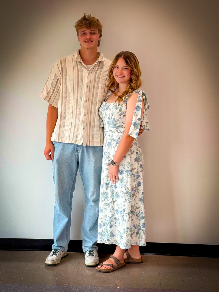 Two students stand side by side indoors against a plain light-colored wall for a posed school portrait. The student on the left is wearing a light striped short-sleeved button-up shirt, light blue jeans, and white sneakers. The student on the right is wearing a white ankle-length dress with a blue floral pattern, shoulder-tie straps, a smartwatch, and brown sandals. Both are facing the camera and standing close together on a tiled floor, with a black baseboard visible along the wall behind them.