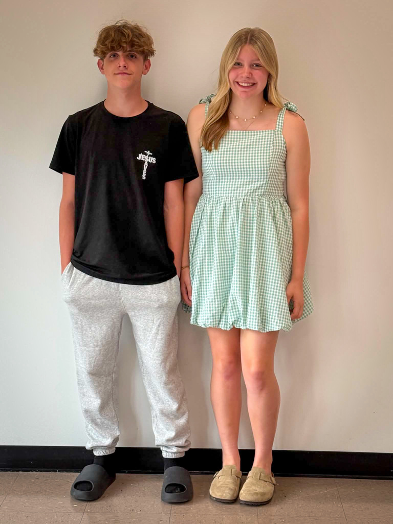 Two students stand side by side in front of a plain light-colored wall for a posed school portrait. The student on the left is wearing a black T-shirt with white lettering on the chest, light gray sweatpants, and dark slide sandals. The student on the right is wearing a sleeveless light green and white gingham-pattern dress and tan slip-on shoes. Both are facing the camera with their arms at their sides. The photo is taken indoors with a tiled floor visible at the bottom of the image.