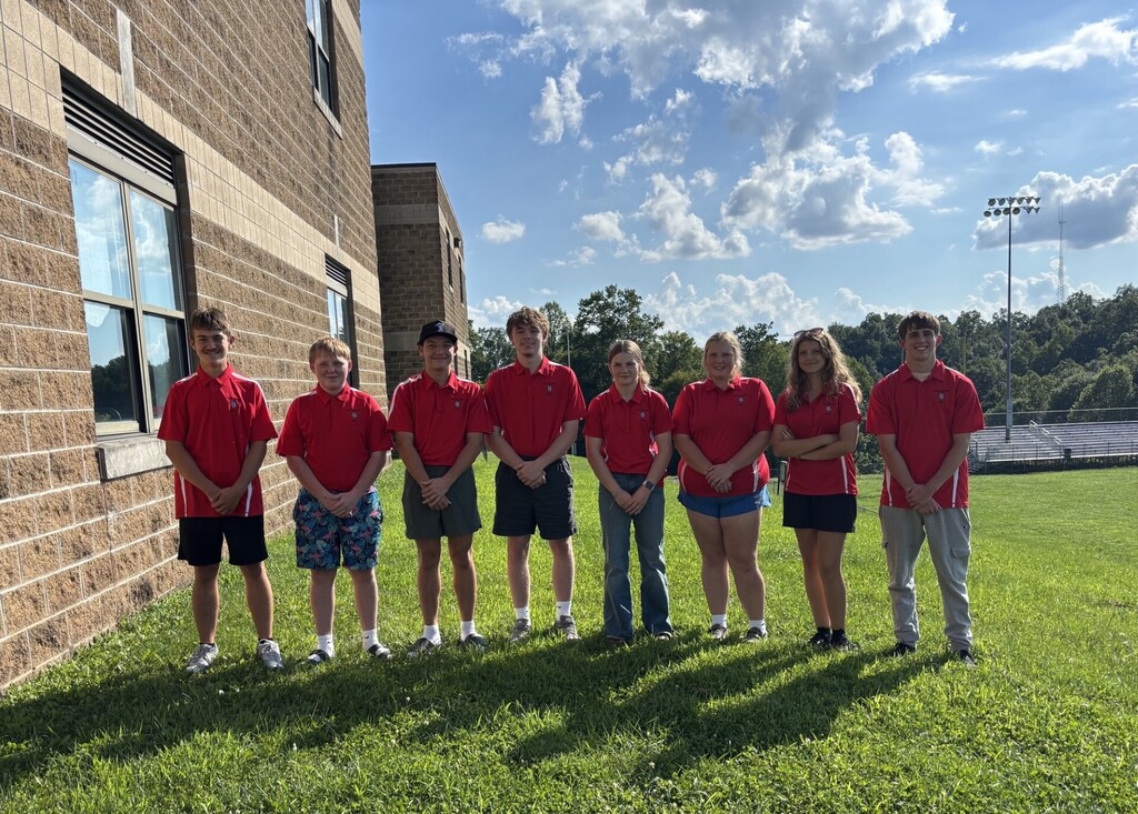 Eight students wearing matching red polo shirts stand in a line on a grassy area beside a school building. The group is posing outdoors on a sunny day under a blue sky with scattered clouds. Trees and athletic field bleachers are visible in the background. Students wear a mix of shorts, jeans, athletic shoes, sandals, and boots. The photo appears to be a team portrait taken near the school campus, with the brick exterior of the building visible on the left side of the image.