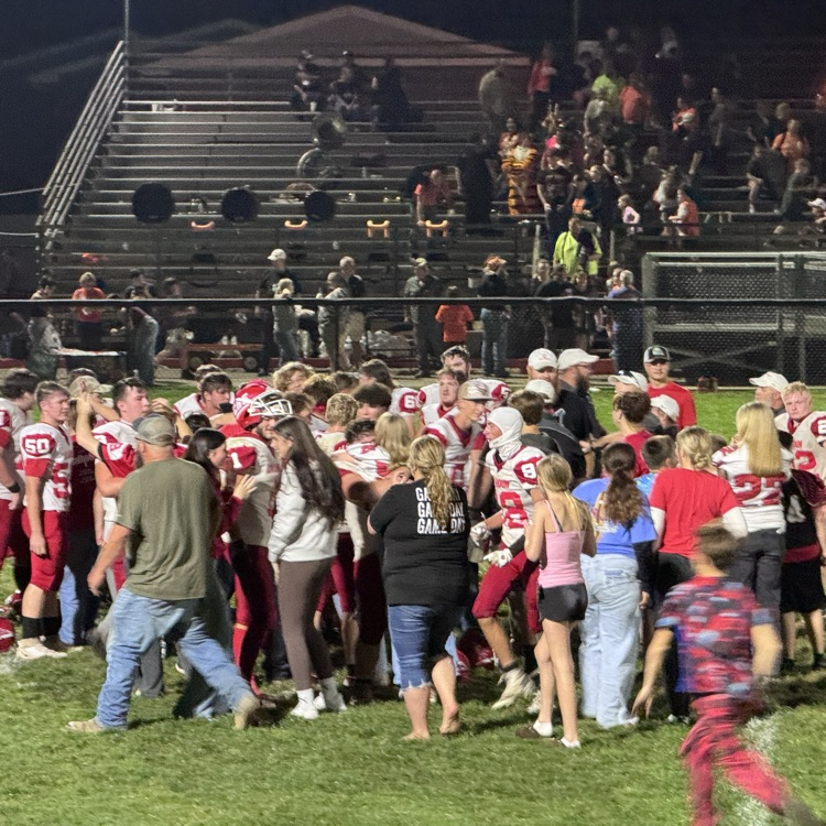 football team celebrate on the field in a circle. fans join on the field to celebrate the win