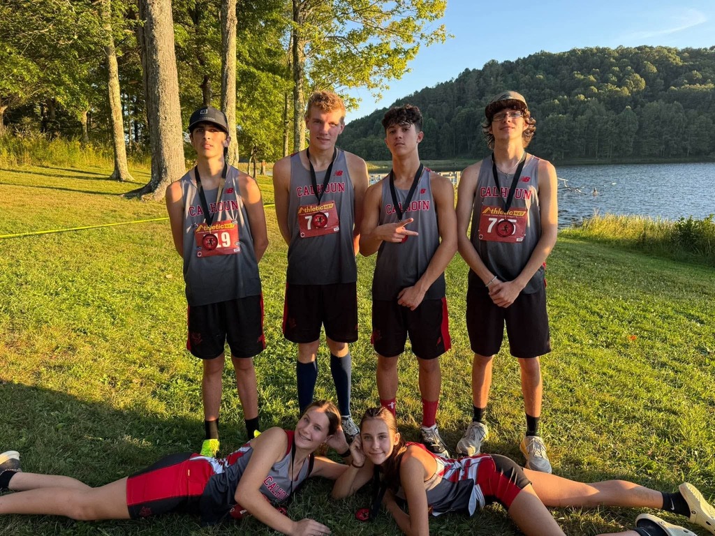 Teammates pose with winnings from the meet.  Four boys standing  behind two girls who are laying on teh ground vertically looking at camera. 