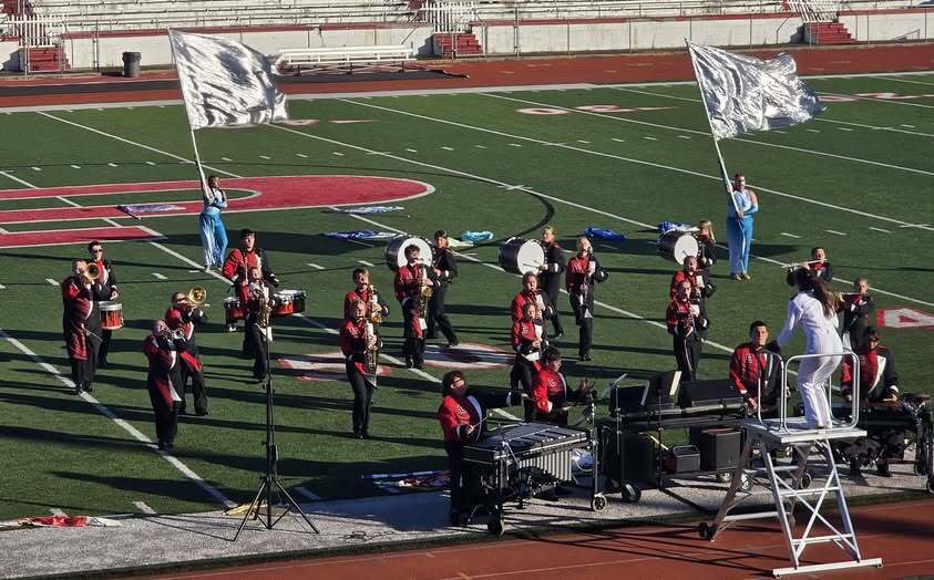 Calhoun County High School Marching Band performs on a football field during the Jim Kessel Invitational on September 26, 2026. Band members in red and black uniforms are arranged in formation with percussion, brass, and woodwind sections visible. A drum major conducts from a podium near the front of the ensemble while two color guard members display silver flags.