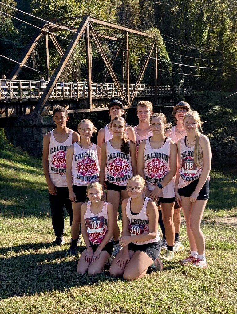 Nine Calhoun Red Devils athletes pose together outdoors in front of a metal truss bridge.