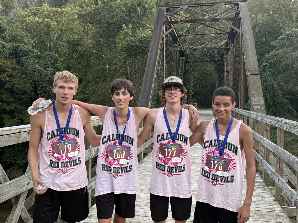 Four Calhoun Red Devils athletes in pink shirts and medals stand on a wooden bridge surrounded by greenery.