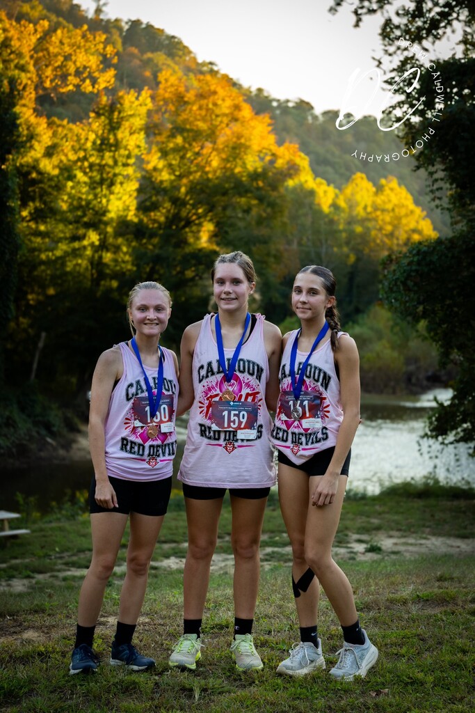Three Calhoun Red Devils runners wearing medals stand outdoors with fall trees and a creek behind them.