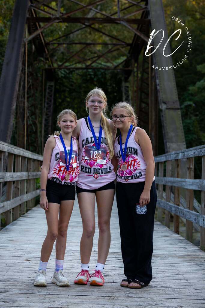 Three Calhoun Red Devils athletes stand on a wooden bridge wearing pink shirts and medals.