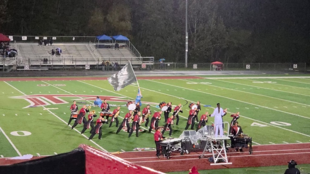 Band preforms on green turf field, solo silver flag in the air. Preforming band members are leaning to the right. 