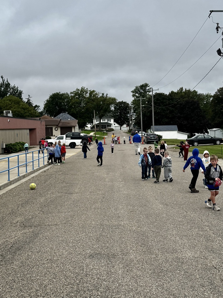 Elementary students play with balls and gather with friends on the paved area outside the school while adults supervise.