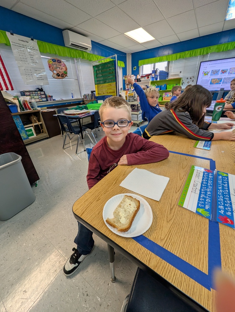 Student at table with his homemade bread in front of him on desk. Button on the bread. Students in the background.