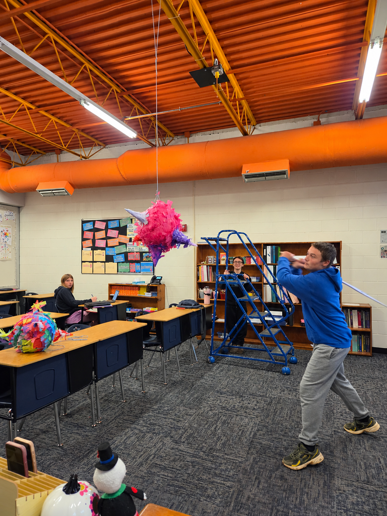 A student swings a stick toward a hanging pink and purple piñata while classmates watch in Spanish Culture class.