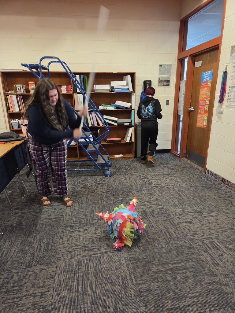 A student raises a stick to strike a colorful handmade piñata resting on the classroom floor.