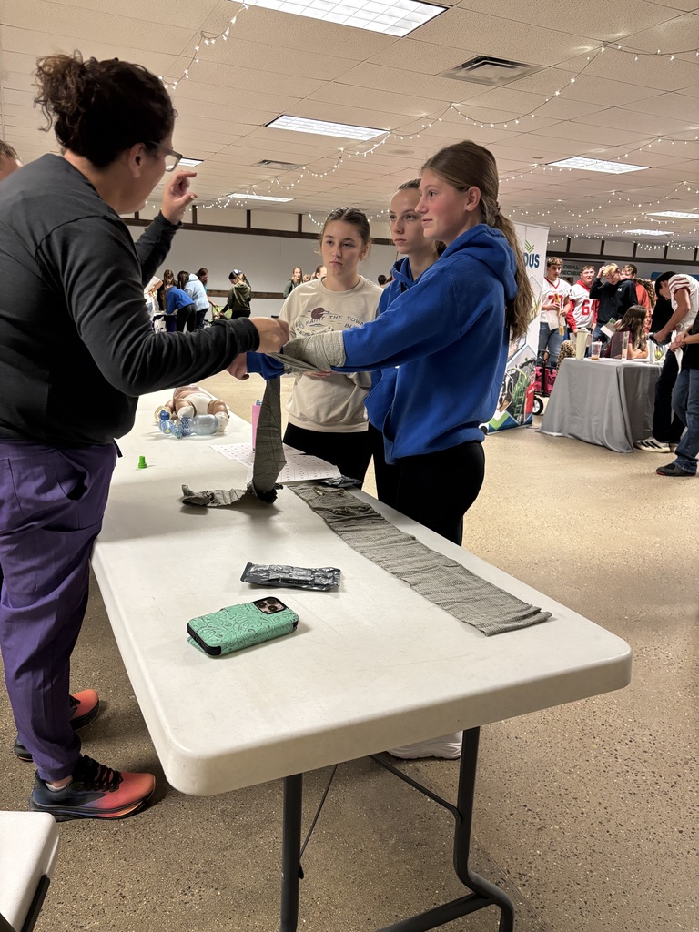 A presenter demonstrates wrapping a student’s forearm with a bandage while two other students watch.