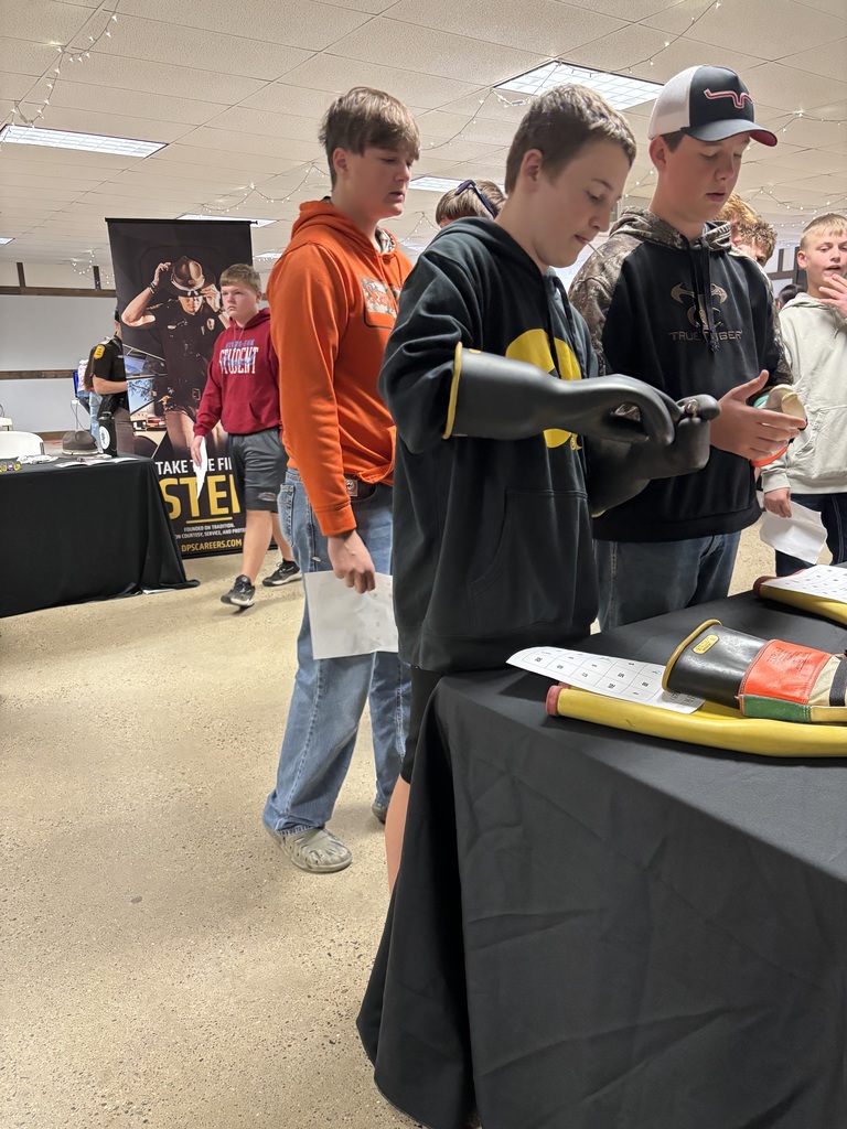 Students try on large protective rubber gloves at a career fair booth with safety equipment on display.