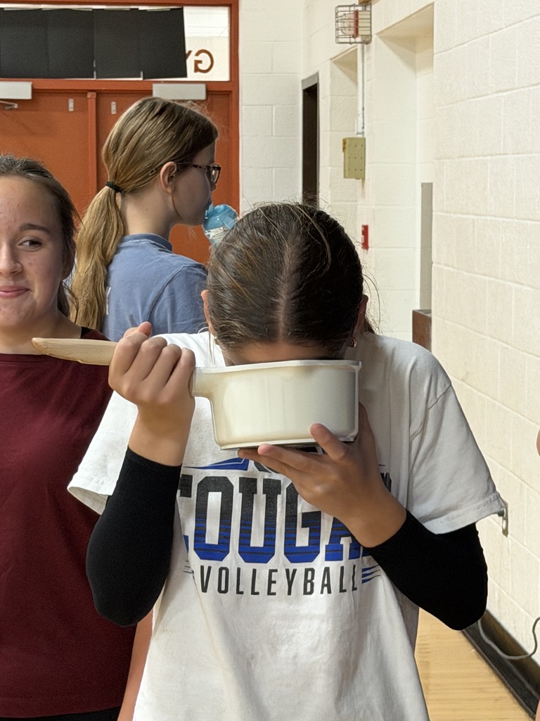 one volleyball player drinking out of a sauce pan with one girl watching and smiling and another drinking out of a sippy cup in the background