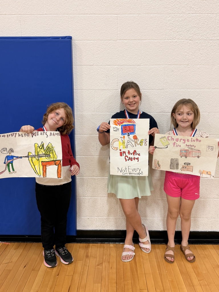Three CAM students wearing medals display handmade fire safety posters featuring a firefighter, a burning battery, and a fire truck.