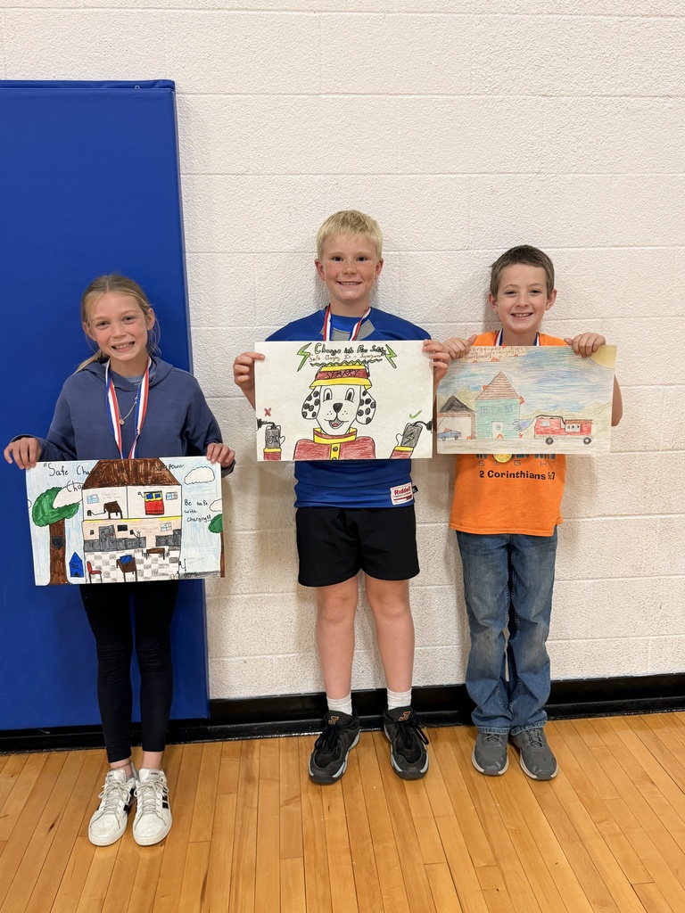 Three CAM students wearing medals display fire safety posters featuring safe charging at home, Sparky the Fire Dog, and a fire truck.