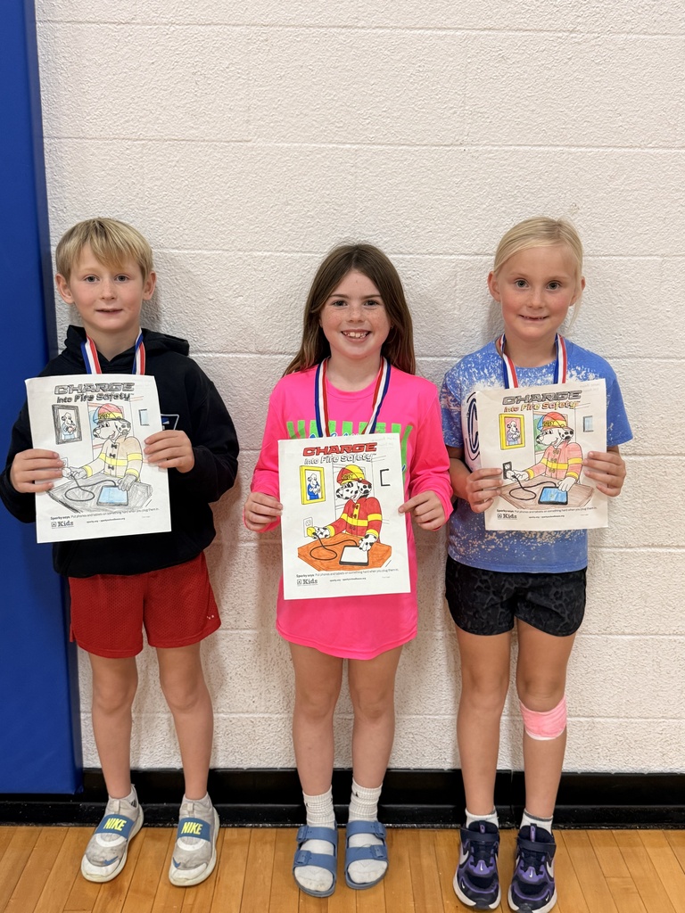 Three CAM students wearing medals hold “Charge into Fire Safety” coloring pages showing Sparky the Fire Dog plugging in a device.