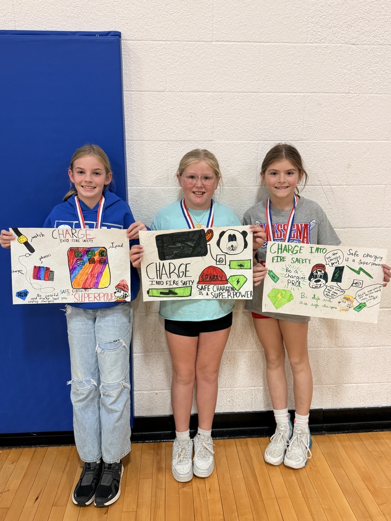 Three CAM students wearing medals hold colorful “Charge into Fire Safety” posters featuring devices, batteries, and messages about safe charging.