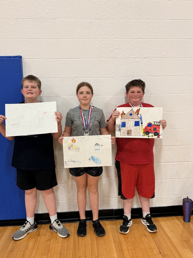 Three CAM students wearing medals hold handmade fire prevention posters showing charging safety examples and a fire truck beside a burning house.