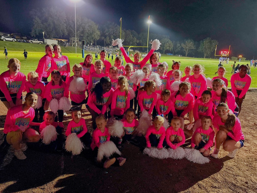 Group of cheerleaders and youth cheerleaders smiling in neon pink shirts with white poms gathered together in a group