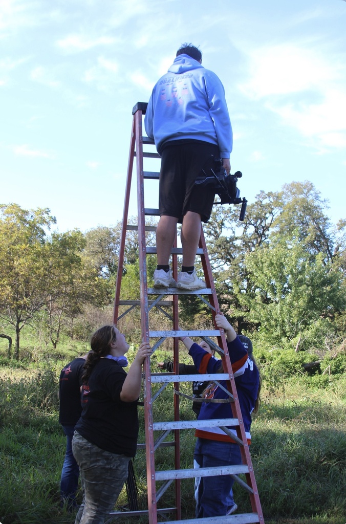 A student holds a video camera while standing on a ladder outdoors as classmates steady the ladder below.