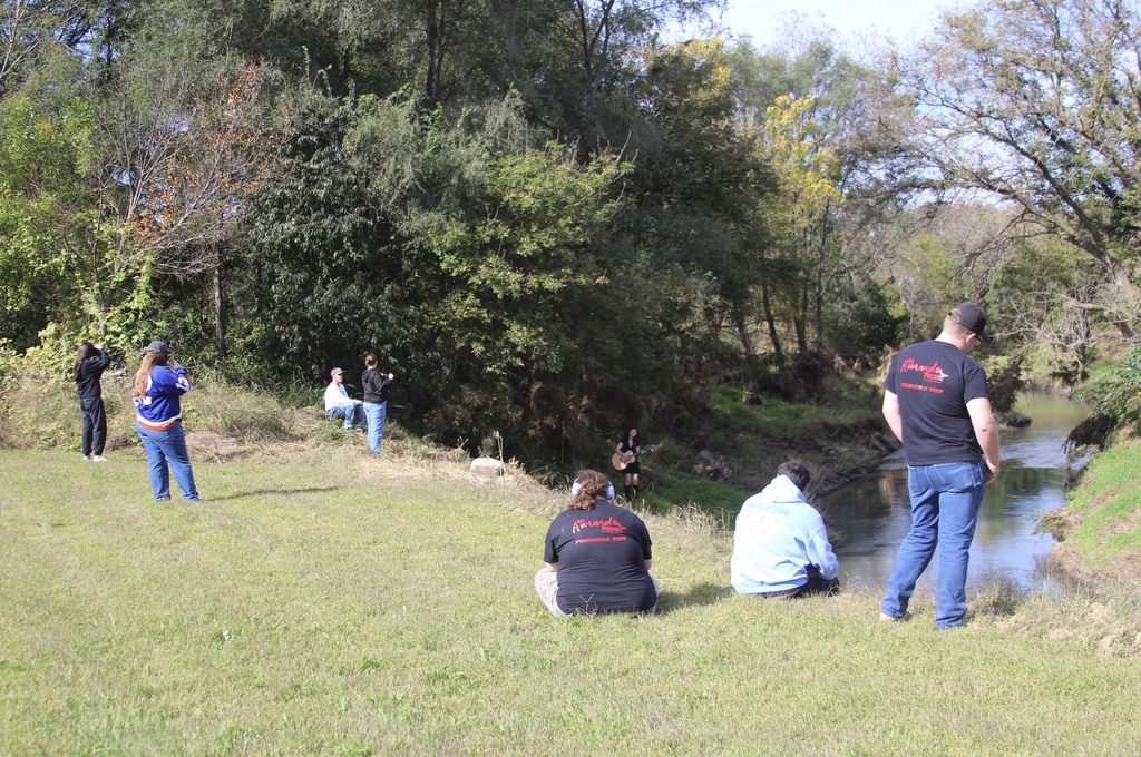Students spread out along a grassy creek bank during a music video shoot as a musician plays an acoustic guitar near the water.