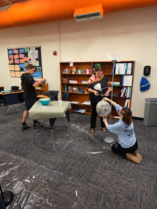 Students work on a newspaper-covered papier-mâché balloon for a homemade piñata, with supplies on a nearby table.