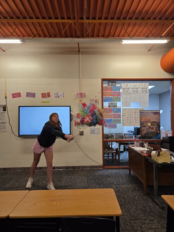 A student swings a stick at a colorful hanging piñata in the Spanish classroom.