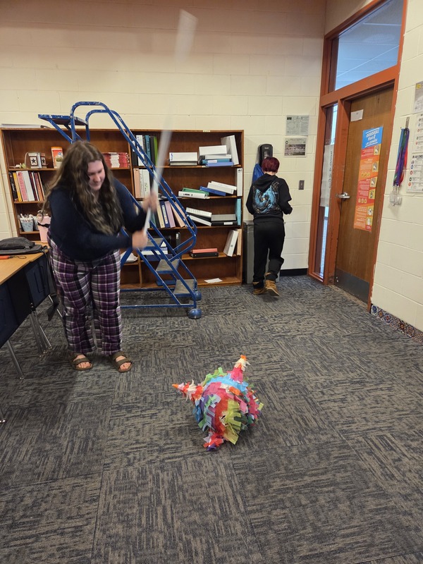 A student raises a stick to strike a colorful handmade piñata resting on the classroom floor.