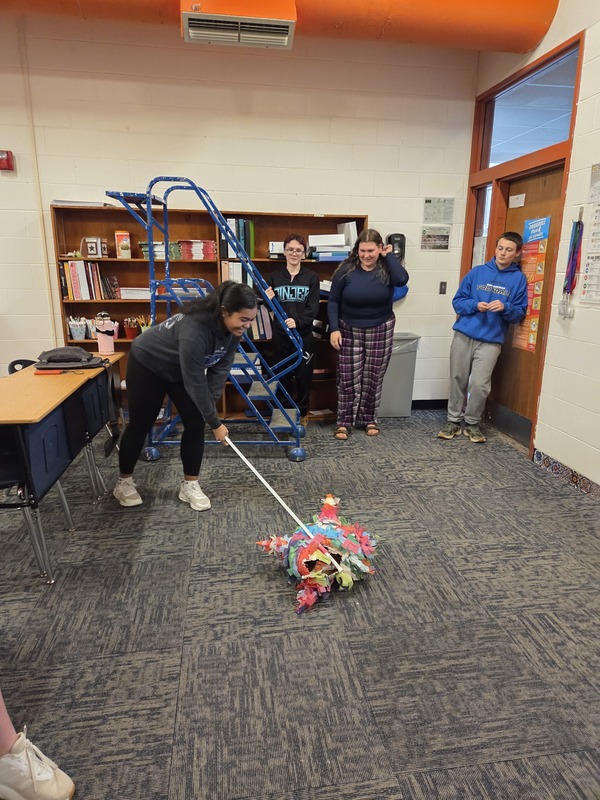 A smiling student uses a stick to break open a colorful piñata on the classroom floor while three classmates watch.
