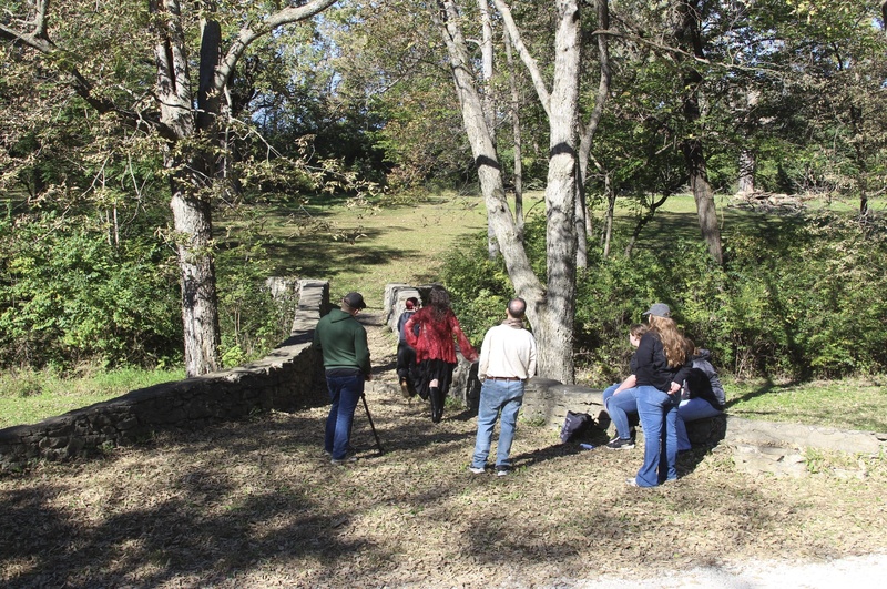 Students and production crew gather on a stone bridge surrounded by trees, with a camera tripod set up for the music video shoot.