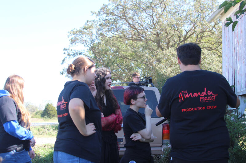 Students and a performer gather beside a pickup truck and wooden building during the music video shoot. Several students wear “The Amanda Project” production crew shirts.